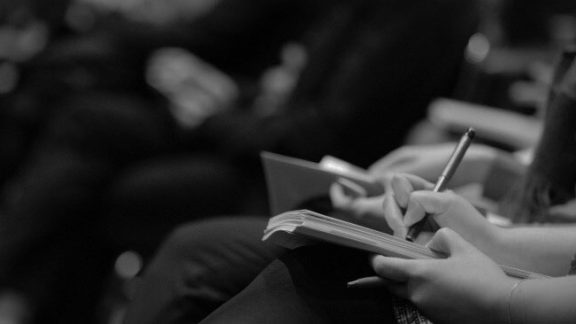 Person writing in their notebook, seated in a row with others who are blurred.