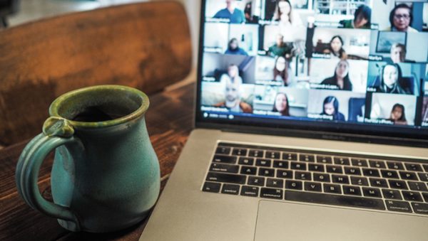 mug on wooden desk beside laptop showing online meeting with 16 people.