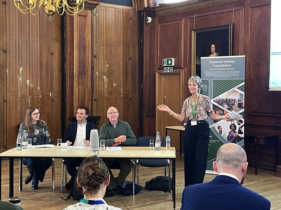 A woman stood addressing an audience, with three people seated in a panel behind a table.