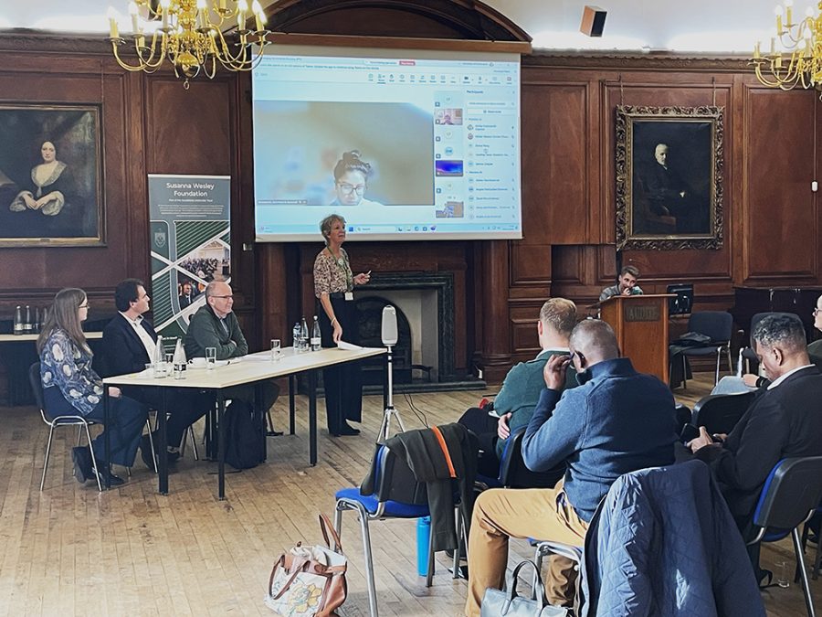 Panel of 3 and standing woman addressing audience, with another person joining online on a screen behind.