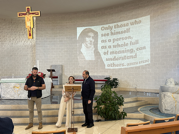 Three people on a stage near a microphone. Background shows image of Edith Stein and reads 'Only those who see himself as a person, as a whole full of meaing, can understand others.'