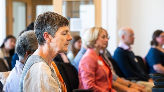 Close up, side-on photo of a woman with short hair, eyes closed, deep in thought. She is in a conference environment and other participants are in the out-of-focus background.