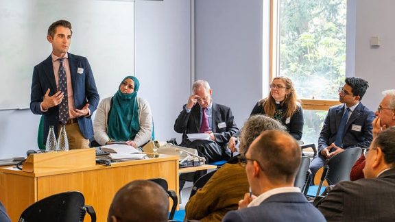 A classroom environment with a man in blue blazer and beige trousers standing and talking to the others. Alongside are a woman in hijab, a man in a suit, a woman with a clerical collar. Photo is taken over the heads of other participants.