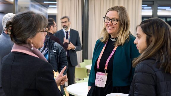 A group of people stand talking indoors, near some tables. A woman wearing a clerical collar and a lanyard saying "Nicola" is listening to another woman in a group of three. the third woman is also watching the speaker. Other adults are in the background.